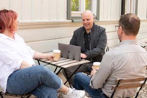 Tre av Midagons medarbetare sitter ner vid ett cafébord utomhus. En har sin dator framför sig och de andra två har varsin kaffemugg i handen.